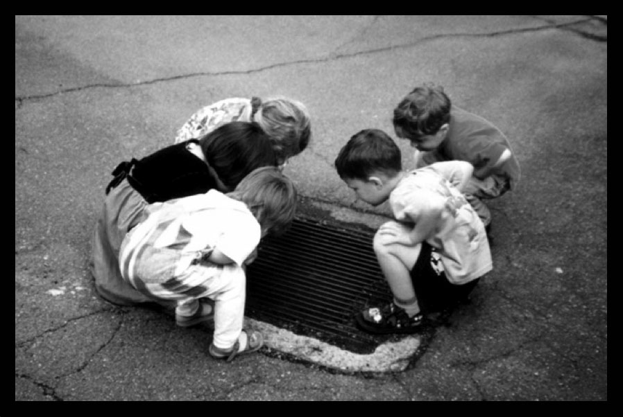 five young children squatting down together to look down into a storm drain